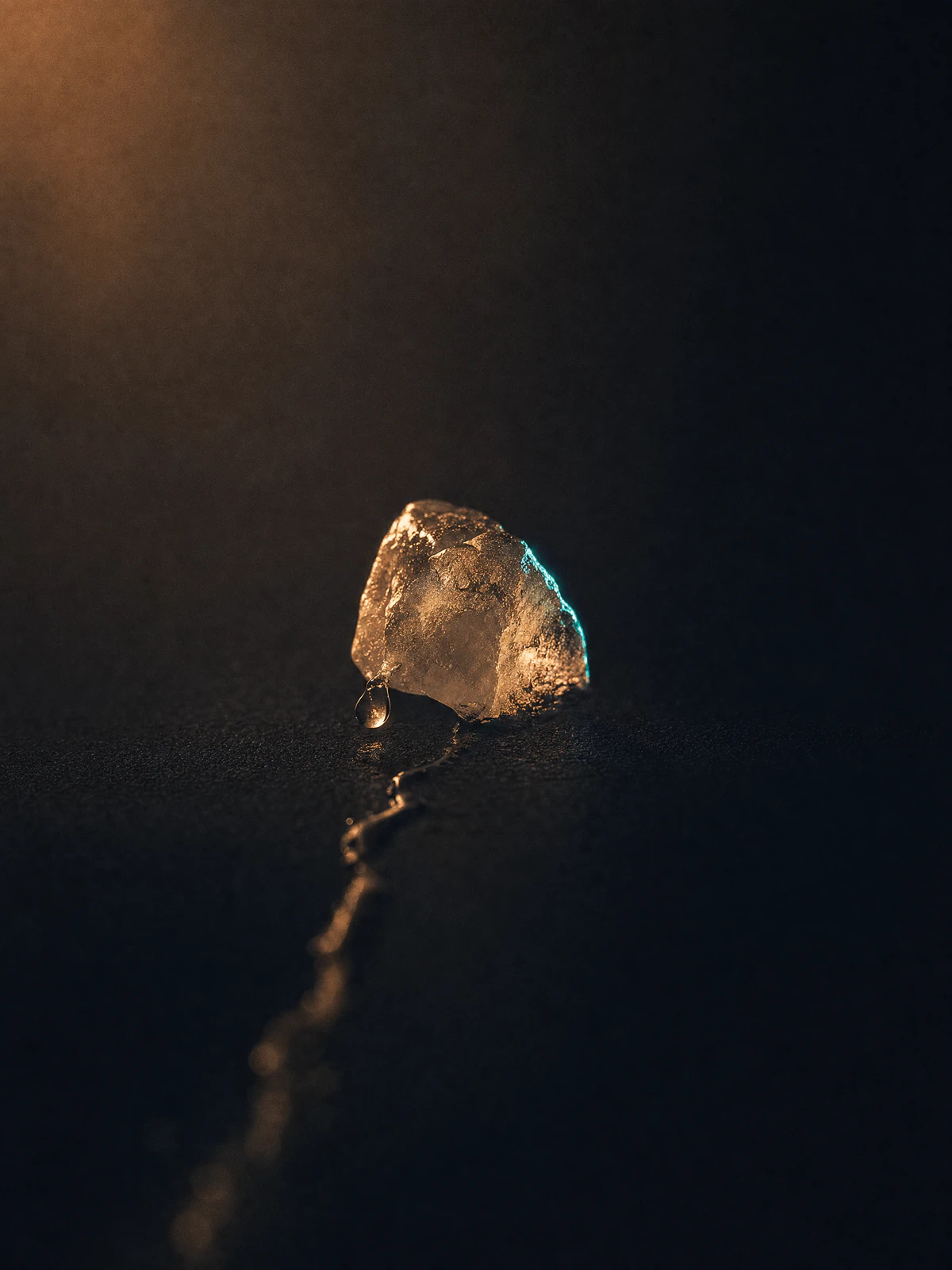 A single ice cube caught mid-melt, already half gone, one bright rivulet running away from it.